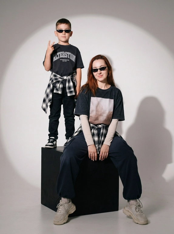 Photorealistic studio portrait photography in minimalistic fashion/editorial aesthetics, static camera at chest level of a seated person, light frontal angle without noticeable perspective deformation; composition with two characters: an model sits on a large matte black cube, body slightly turned towards the camera, knees bent, feet on the floor, the arms are lowered and relaxed in front of the knees, the fingers are neatly extended; the child stands on the same cube behind and to the left, at full height, one hand raised with a "V" gesture, the other lowered, pose confident and playful; both wearing narrow dark sunglasses, gaze directed at the camera; shooting plan — full height for both with a small supply of air above and below, center the center of the frame is shifted to the right to a sitting figure, a standing child balances on the left; the lens is normal/short portrait 50-70 mm, without width; aperture is approximately f/5.6–f/8, moderate depth of field, both characters are sharp, the background is slightly soft; the studio lighting is soft, with one key source on the front left at a level just above the head (softbox), giving smooth transitions and a pronounced but soft shadow against the background on the right; light filling light from the front reduces contrast, shadows are readable; the background is a smooth, light, seamless cyclorama/wall with a noticeable large oval light spot (gradient) in the center, there is a darker vignetting around, a clean minimalistic studio without props; the mood is calm, stylish, slightly retro, with an emphasis on street fashion and family vibe; the expression on the face of the seated model is a soft smile, relaxation; the person standing has a confident neutral facial expression; clothes: the seated model has a dark oversized T-shirt with a large rectangular print on the chest, under it a light longsleeve with long sleeves, wide dark cargo trousers/joggers with pleats at the knees and bottom, a plaid shirt/flannel tied at the waist with a knot, massive light sneakers with a pronounced sole and mesh texture; the child has a dark T-shirt with a large inscription on the chest, dark trousers, a plaid shirt tied at the waist, sneakers in two colors; fabrics with a realistic cotton and flannel texture, noticeable fine folds, natural creases on the knees and in the waist area, matte surfaces without gloss; micro-details: natural skin texture without plastic, neat glare on the lenses of glasses, a clear contour of the shadow from the raised hand on the background, clean floor and soft drop shadow from the cube; high detail, natural sharpness, light grain like a film shot, moderate contrast, soft matte/film look, no HDR, no overexposure and no unnecessary objects.

Какого цвета будут рубашки на поясе?: Чёрно-белый.