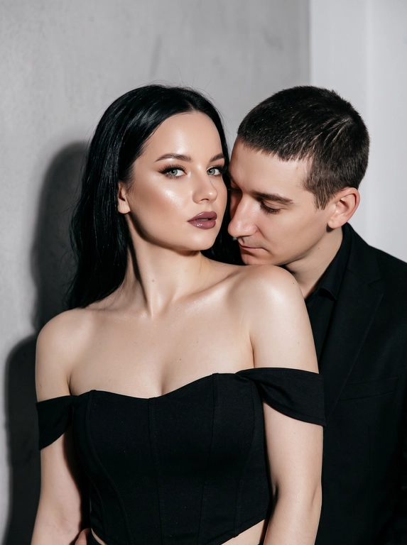 Не меняя черты лица, студийная fashion-съемка пары в минималистичном интерьере с серо-белым фоном, атмосфера гламура и интимной близости, ракурс — крупный портрет по грудь с легким поворотом корпуса, женщина на переднем плане, мужчина позади склоняется к её плечу, поза — она слегка отвернута от камеры с прямым взглядом, он касается лицом её шеи/плеча, одежда — на женщине черный корсет/бандо с открытыми плечами, на мужчине классический черный пиджак и рубашка, макияж — glam (ровный сияющий тон, сильный хайлайтер, четкий контуринг, стрелки, густые ресницы, глянцевые губы), свет — жесткая фронтальная вспышка + мягкий заполняющий свет, выраженные тени позади, эффект flash photography / editorial, холодно-нейтральная цветокоррекция с контрастом и глянцем кожи, высокая детализация, slight film grain, shallow depth of field, съемка на Canon EOS R / 50mm, f/2.0, direct flash, studio look.