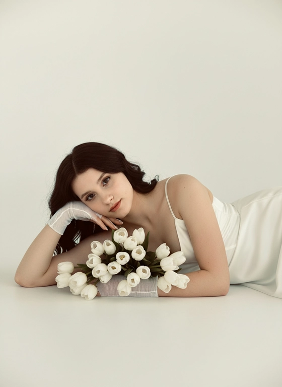 Эта женщина лежит на белой поверхности на боку, голова на согнутой руке. Не меняй ее черты лица, возраст, волосы. Крупный план. На ней молочное атласное платье на тонких бретельках и полупрозрачные белые сетчатые перчатки. Перед лицом лежит букет белых тюльпанов, цветы на переднем плане слегка размыты. Камера на уровне глаз, крупный портрет. Взгляд в камеру, спокойное выражение лица. Мягкий рассеянный дневной свет, белая студия, чистый белый фон. Реалистично, высокая детализация кожи, shallow depth of field.
