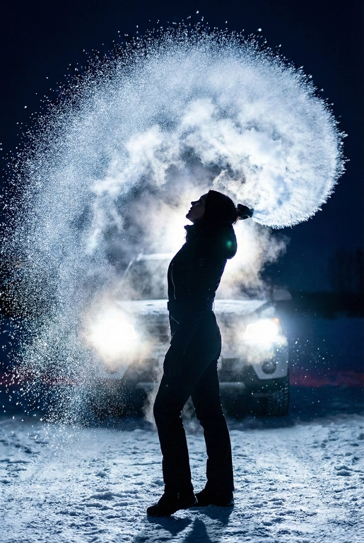 Создай портрет, не меняя черты лица. Эстетичный
женский силуэт в профиль на фоне ночного
снежного пейзажа. Девушка одета в стильный
черный комбинезон. Она в сильный мороз выливает
вверх полукругом в воздух кипяток, который,
замерзая в воздухе, образует красивую огромную
светящуюся дугу в воздухе. Контровой свет от фар
автомобиля сзади создает эффект ореола и
подсвечивает каждую снежинку и пар.
Атмосфера загадочная, холодная, высокая мода,
кинематографичное освещение, гиперреализм,