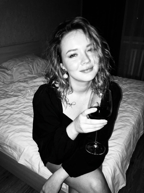 Create a portrait, transfer the face from the reference photo without changing facial features, proportions, or expression, preserve full identity. Aesthetic black-and-white nighttime Y2K photograph captured with direct on-camera flash, shot in motion.
A woman is sitting on the edge of a white, slightly rumpled bed, the frame taken from a slightly elevated angle. She wears a stylish black top with voluminous sleeves and large round statement earrings. In one hand she holds an elegant glass of dark wine. Her body is subtly moving, creating natural motion blur across the face and shoulders, as if the moment was caught mid-gesture. The gaze is languid and directed toward the camera, with a soft, intimate smile. Hair falls in large waves with loose strands drifting across the face, slightly displaced by movement. Lighting is harsh flash against deep night shadows, high contrast with blown highlights and strong falloff, visible film grain. Skin texture remains real and alive, no smoothing, enhanced by flash glare. The mood is high-fashion editorial, Parisian chic, old-money sensuality, nighttime melancholy, spontaneous and cinematic, shot in a raw 35mm film aesthetic with pronounced motion blur and lived-in realism.