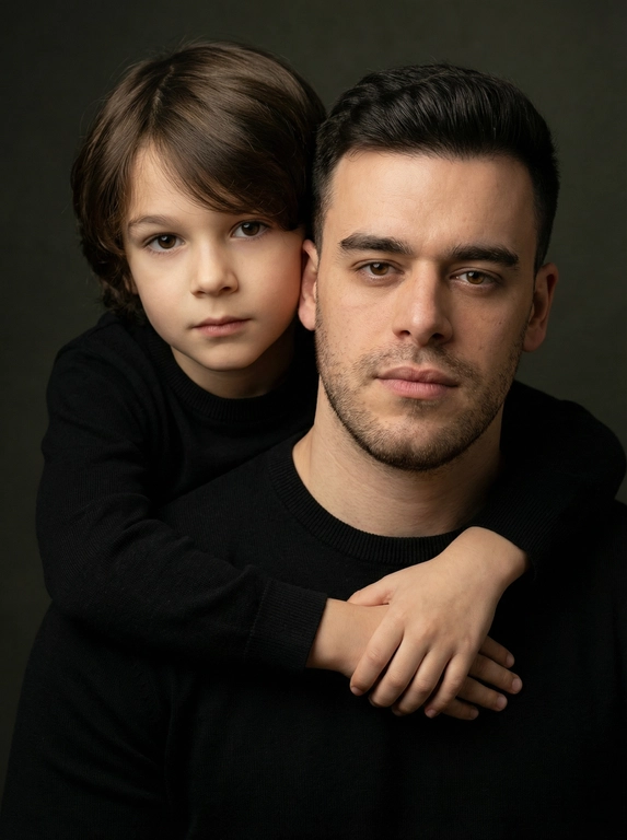 Создай портретный студийный кадр из фотосессии отца и сына. Сын обнимает отца сзади, лежа подбородком на его плече, руки мягко сомкнуты на груди мужчины. Оба смотрят прямо в камеру. Естественные спокойные эмоции, лёгкая серьёзность во взгляде.
Одежда — однотонные чёрные лонгсливы или свитера без принтов. Тёмный нейтральный фон (глубокий серо-оливковый или графитовый). Мягкий направленный студийный свет с лёгкой теневой глубиной, акцент на лицах, тёплая естественная цветокоррекция, высокая детализация кожи, натуральная текстура без излишней ретуши.
Кадр — крупный или средний план, плотная композиция, лица расположены близко друг к другу. Атмосфера — тёплая, камерная, эмоциональная, ощущение близости и доверия. Профессиональная съёмка, 85mm lens, shallow depth of field, cinematic portrait, ultra realistic.

