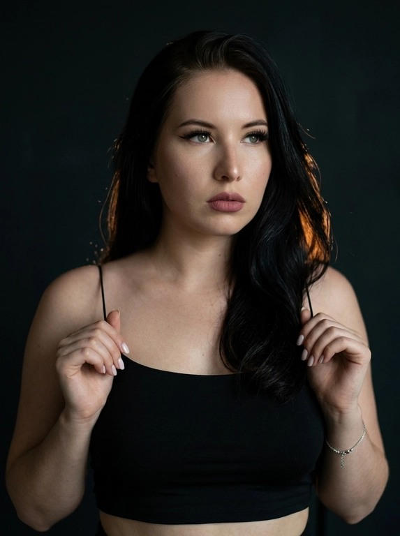 Максимально реалистичная фотография в стиле moody boudoir editorial, передающая атмосферу интимности, чувственности и приглушенного драматизма.
Крупный план по пояс (waist-up shot / close-up). женщина позирует в полумраке, придерживая руками края одежды. [Поза/взгляд/эмоция]: Плечи слегка приподняты, руки с длинным светлым маникюром сжимают тонкие бретели черного топа; голова немного повернута в сторону, взгляд задумчивый и отведен в сторону; выражение лица томное, загадочное и уверенное.
Вечерний «glam» выразительные стрелки, густо накрашенные ресницы, создающие эффект «кошачьего взгляда», мягкий контуринг лица и матовая помада цвета пыльной розы.
Облегающий черный кроп-топ на очень тонких бретелях-спагетти. Материал — матовая эластичная ткань. На запястье виден изящный серебряный браслет.
Минималистичный интерьер в темных тонах. Фон однотонный, слегка текстурированный, уходящий в глубокую тень, что создает эффект камерности.
 Низкий ключ (low-key lighting). Мягкий, теплый направленный свет падает сбоку, подчеркивая рельеф ключиц, линию плеч и текстуру кожи. Глубокие, кинематографичные тени создают объем и драматичное настроение.
 85mm f/1.4, идеальное фокусное расстояние для портрета, создающее минимальные искажения и сильное размытие фона. Очень малая глубина резкости; фокус идеально наведен на глаза и лицо модели, в то время как плечи и руки уходят в мягкое расфокусирование.
Гиперреалистичная текстура кожи: видны поры, естественный блеск на выступающих частях лица и плеч. Отсутствие «пластикового» эффекта ретуши.
 текстура ткани топа под пальцами, тонкие волоски на макушке, мягкий градиент тени на стене.
Эстетика «dark feminine energy», интимная, теплая и дорогая атмосфера (expensive moody vibe).

Какая будет прическа?: Волосы: перекинутые на одно плечо. Текстура волос гладкая и шелковистая, с отдельными тонкими прядями, подсвеченными теплым светом.