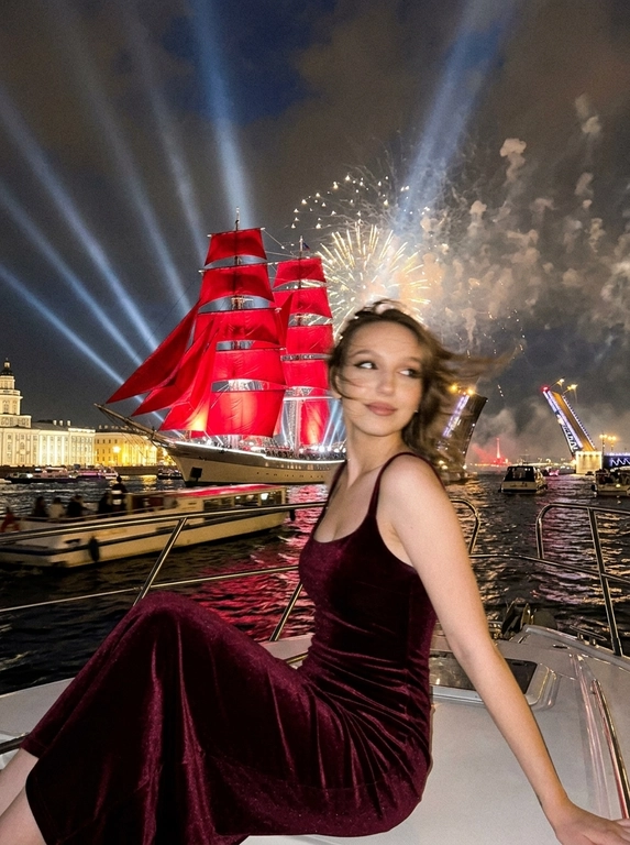 Создай портрет. Ночной экстремально близкий портрет в стиле дорогого fashion-снимка, крупный план, ощущение случайно пойманного момента. Модель, сидящая в энергичной позе на роскошной лодке, плывущей по каналам Санкт-Петербурга во время праздника - "Алые паруса". Волосы развиваются от ветра, слегка на лице. Шикарный вечерний макияж, у нее еле заметная улыбка. 

На заднем плане воды канала, отражающие на горизонте появляются исторические здания, характерные для Санкт-петербурга, рядом с лодкой плывет огромное количество парусных лодок, маленьких яхт, туристических лодок и небольшие лодки вдалеке. Интерьер лодки изысканный. На фоне разводной мост и в центре внимания огромный известный корабль с алыми парусами, паруса подсвечены, с корабля красиво бью прожектора, он в центре внимания, выглядит величественно. Здания подсвечиваются. Все небо залито огромным количеством салютов и фейерверков, а так же светом прожекторов, салюты во весь кадр

Кадр намеренно неидеальный: сильный motion blur, длинная выдержка, высокий ISO, выраженное зерно. Жёсткий контраст, кинематографичная небрежность, ощущение ночи, роскоши и живого, спонтанного кадра. Атмосфера праздника, величественного события. Модель сидит грациозно прямо к кадру, голова повернута в сторону, лицо немного размазано из-за движения, девушка в расфокусе, не смотрит в кадр. Весь фокус на корабле с алыми парусами, они четко подсвечены.

Атласная красная лента "Выпускница 2026"?: Без ленты

Какое будет платье?: Бордовое вечернее платье, винный оттенок, гладкий атлас или бархат, приталенный силуэт, длинная юбка, возможна мягкая драпировка на талии. Образ эффектный, уверенный, немного драматичный.