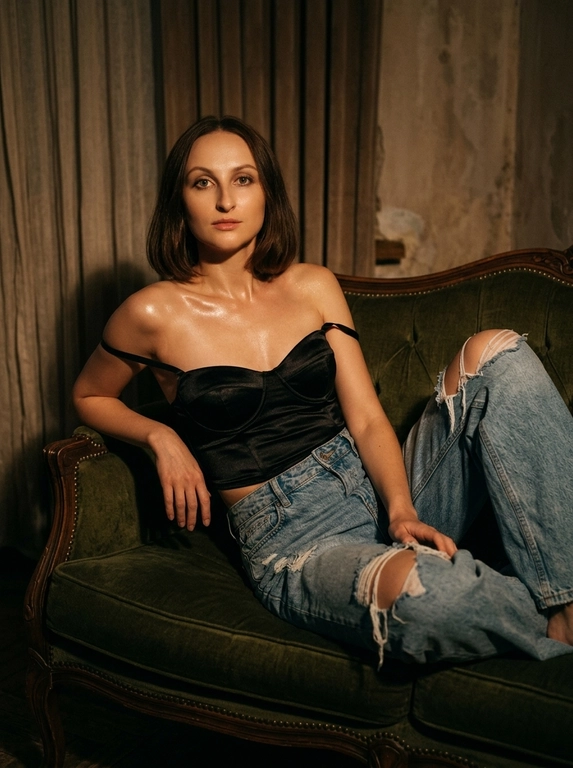 Абсолютное сохранение черт лица модели, внешность 1:1, не искажать и не менять лицо.
Общая стилистика:
Глубокий, атмосферный, интимный fashion editorial портрет с кинематографическим качеством. Изображение должно быть "moody" (настроенческим), с высоким контрастом и ощущением пленочной фотографии.
Поза и Ракурс:
Женщина в расслабленной позе полулёжа откинулась назад на винтажном бархатном диване глубокого цвета. Корпус развернут в 3/4, одна рука служит опорой за спиной, ноги согнуты в коленях и перекрещены. Ракурс камеры чуть ниже уровня глаз, создавая ощущение подглядывания. Кадрирование по бедра (3/4 роста), основной акцент на ключицы, плечи и зону декольте.
Внешность и Одежда (Без указания цвета волос):
Макияж: Акцент на длинные, разделенные черные ресницы. Мягкий скульптурирующий контуринг лица, естественный румянец. Заметный влажный хайлайт на ключицах и плечах.
Освещение и Интерьер:
Свет: Драматичное, теплое боковое освещение (как от окна на закате) служит рисующим светом, создавая глубокие тени и подчеркивая объем. Тонкий, слабый контровой свет (rim light) едва очерчивает силуэт плеч и прически, отделяя модель от фона.
Окружение: Комната в полумраке. На фоне угадываются тяжелые фактурные портьеры, старая штукатурка на стене или деревянные панели. Атмосфера будуара или старой квартиры.
Технические и Художественные Детали:
Эстетика пленочного кадра, снятого на объектив 35mm или 50mm с открытой диафрагмой (f/2.8), обеспечивающей мягкое, художественное боке на заднем плане. Изображение слегка недоэкспонировано (low key) для насыщенности теней. Цветокоррекция в тёплых, приглушённых бежево-коричневых и охристых тонах с пониженной насыщенностью холодных оттенков (зеленого и синего). Высокая детализация текстуры кожи, ткани денима и сатина, подчёркнутая легкой, естественной пленочной зернистостью и мягкой виньеткой по краям кадра.

Какая будет прическа?: Не изменять мою прическу.

Какая будет одежда?: Черный корсет-бюстье из шелковистого сатина с приспущенными бретелями. Светлые, свободные джинсы из грубого, сильно рваного и потертого денима.