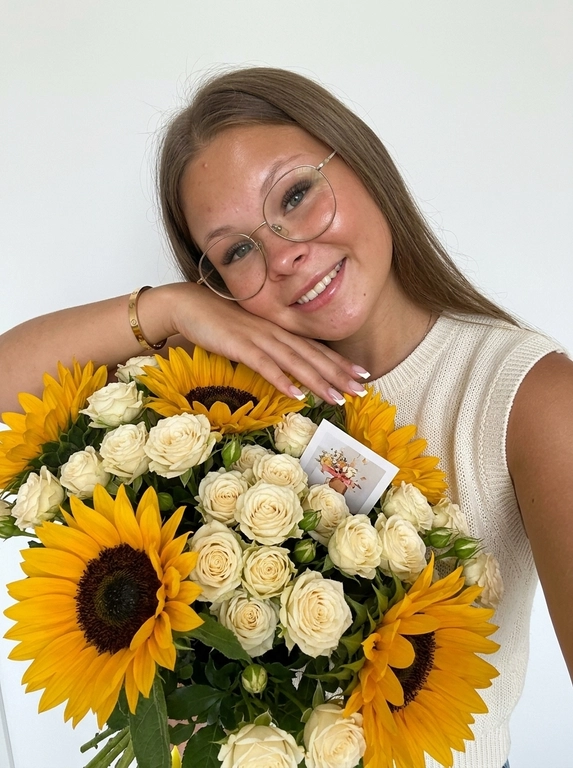 На фотографии запечатлена женщина, которая держит в руках очень большой и пышный букет цветов.
Её кожа сияет, загорелого оттенка, на губах глянцевый блеск, улыбка лёгкая.

она положила руку и голову на букет, взгляд в камеру и находится позади букета.
 Букет: Это объемная композиция, состоящая из контрастного сочетания двух видов цветов:
    – Подсолнухи: Крупные, яркие желтые подсолнухи с темной сердцевиной, которые создают акцент в букете.
    – Розы: Множество нежных кремово-белых роз (похожих на кустовые), которые заполняют пространство между подсолнухами, придавая композиции мягкость и элегантность.Среди цветов видна небольшая подарочная карточка или визитка.

Она одета в вязаный топ с высоким горлом, без рукавов, свет молочный. Маникюр аккуратный, Френч, квадратная форма, на запястье браслет Cartier. Композиция выглядит очень ярко, позитивно и празднично благодаря сочетанию солнечных оттенков подсолнухов и нежных тонов роз.

Фон - белая стена. Высокая детализация кожи. Живое фото, без ретуши на лице. Мобильная съемка. Лёгкая пленка, зерно. Raw. ультрареалистичный дорогой лайфстайл снимок.
