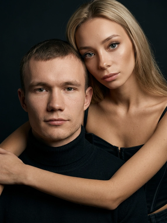 Гиперреалистичный студийный fashion-портрет молодой пары по загруженному референсу, 100% сходство лиц. Черты лица не изменять. Крупный план, кадр по плечи. Мужчина, спокойный уверенный взгляд прямо в камеру, естественное выражение лица, одет в черный трикотажный свитер с высоким воротом. Женщина стоит позади мужчины и нежно обнимает его, ее руки лежат на его плечах. Ее лицо расположено рядом с его лицом, она повернута слегка боком, голова немного наклонена вперед, взгляд направлен прямо в камеру. У женщины выразительные глаза с длинными густыми черными ресницами, естественный clean-glam макияж, натуральные брови, теплый оливковый оттенок кожи, небольшие серьги-кольца. Композиция плотная и интимная, их лица расположены очень близко друг к другу, современный модный портрет пары. Женщина одета в черный корсет. Кожа максимально натуральная: живая текстура кожи, видимые микропоры, subtle skin glow, естественный мягкий блеск кожи, natural skin microtexture, skin translucency, micro skin details, легкие естественные несовершенства кожи, без ретуши и без эффекта пластика. Освещение: мягкий студийный beauty lighting, рассеянный ключевой свет спереди и немного сбоку, мягкие глубокие тени для объема лица, деликатные световые блики на коже. Фон: глубокий темный студийный фон, темно-графитовый или почти черный, создающий контраст с кожей и лицами. Стиль: luxury fashion editorial photography, high fashion portrait, ultra photorealistic, 85mm portrait lens, shallow depth of field, creamy bokeh, cinematic soft lighting, RAW photo, extremely detailed, fine hair strands, natural skin pores, professional photography, photorealism, 8k, high dynamic range.