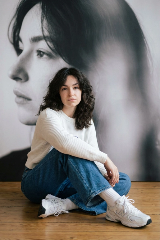   Максимально сохранить внешность, причёску и длину, как на фото.
Женщина должна сидеть на полу в изящной позе, в современном белом джемпере , в широких джинсах и белых носках и белых  кроссовках New Balance.

Её волосы как на фото
Выражение лица, как на фото, она смотрит на камеру .

Фон — тот же её профиль, в мягком крупном чёрно-белом художественном стиле, крупный план сбоку.

Композиция должна быть художественной, мягкой, реалистичной.
Создай изображение, которое выглядит как настоящая фотография.
