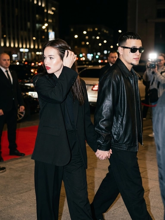 Ultra-realistic nighttime paparazzi-style photograph. A candid shot of a couple walking at night. The woman walks in front, brushing her hair back with one hand, while the man walks slightly behind her at a diagonal angle. They are holding hands. The camera is positioned slightly above eye level with a close zoom.
Preserve the celebrity magazine photography aesthetic exactly.
The faces MUST NOT change. The original facial features, eyes, nose, lips, hairline, and skin texture must remain completely identical. There should be no AI-generated look.

The man is wearing a loose oversized black leather jacket and Ray-Ban sunglasses. Charismatic, wealthy, and stylish look.
The scene is shot at night with a harsh paparazzi flash. In the background, there are blurred city lights and the atmosphere of a luxury venue. The photo should look as if a celebrity couple was secretly photographed by paparazzi, natural yet professionally captured.
Canon EOS R5, 85mm lens, ultra-realistic skin texture, cinematic lighting, natural motion, Vogue and celebrity magazine aesthetic.
IMPORTANT:
* The woman walks in front, the man walks behind.
* The woman appears to be brushing her hair back with one hand.
* The woman and man are holding hands.
* The shot should look as if a paparazzi flash has just fired.
* Do not alter, beautify, or transform the faces into different people.

Выберите одежду для девушки: : The woman is wearing a black old money outfit: relaxed yet extremely elegant, with an oversized black blazer or a loose black top, wide black trousers, and a luxurious minimal appearance. Quiet luxury aesthetic.