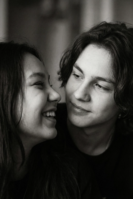   Сгенерируй Черно-белое фото, кинематографичная фотография пары, снятая крупным планом. девушка находится слева в кадре на переднем плане, повернута в профиль и смеется, не глядя на парня , парень справа на заднем фоне смотрит на глаза девушки задумчиво и влюбленно, с мягким, нежным, влюбленным выражением лица. свет падает сбоку, создавая мягкие тени на лицах и подчёркивая черты лица . фон размытый , темный , без четких деталей , создававший атмосферу уюта фотография выглядит как кадр из романтического фильма или личный момент пойманный камерой формат фото