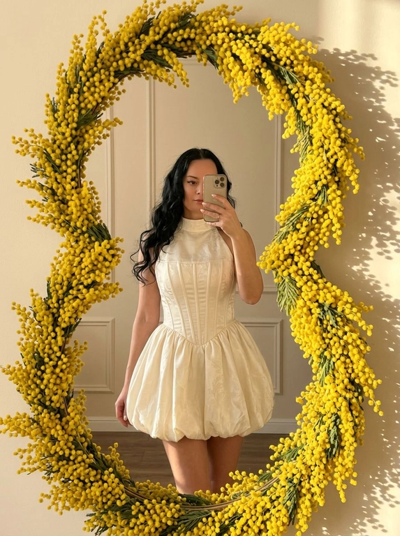Зеркальное селфи в интерьере с мягким тёплым дневным светом. Большое зеркало в форме цифры 8 с плавными округлыми линиями занимает значительную часть кадра. Рама полностью обрамлена густыми ветвями жёлтой мимозы, пушистые цветы создают яркий солнечный контур вокруг отражения. Лёгкие тени от мимозы падают на светлую стену. Кадр в полный рост. Женщина стоит перед зеркалом в лёгком полуобороте, вес перенесён на одну ногу, силуэт вытянутый и уверенный. Телефон держится на уровне лица, частично закрывает одну сторону, но черты остаются читаемыми. Макияж — нюдовый и элегантный: ровный мягкий тон, лёгкое естественное сияние кожи, небольшие растушёванные коричневые стрелки, аккуратные ресницы. Губы с глянцевым прозрачным блеском. Маникюр аккуратный. Свет тёплый, золотистый, с мягким контрастом. Атмосфера весенняя, романтичная, но с акцентом на архитектуру силуэта. 

Какая будет одежда?: Наряд — молочно-кремовое мини-платье с корсетной архитектурой. Верх — высокое закрытое горло с плавным переходом в чётко сконструированный корсетный лиф. Выраженные вертикальные рельефы подчёркивают талию и формируют sculpted силуэт. Ткань плотная, с деликатным жаккардовым цветочным рисунком и лёгким сатиновым свечением. Низ — объёмная пышная юбка-баллон с мягкими складками, создающая контраст между узкой талией и округлой формой низа. Длина мини, подчёркивает линию ног. Силуэт couture inspired, структурированный, но элегантный.