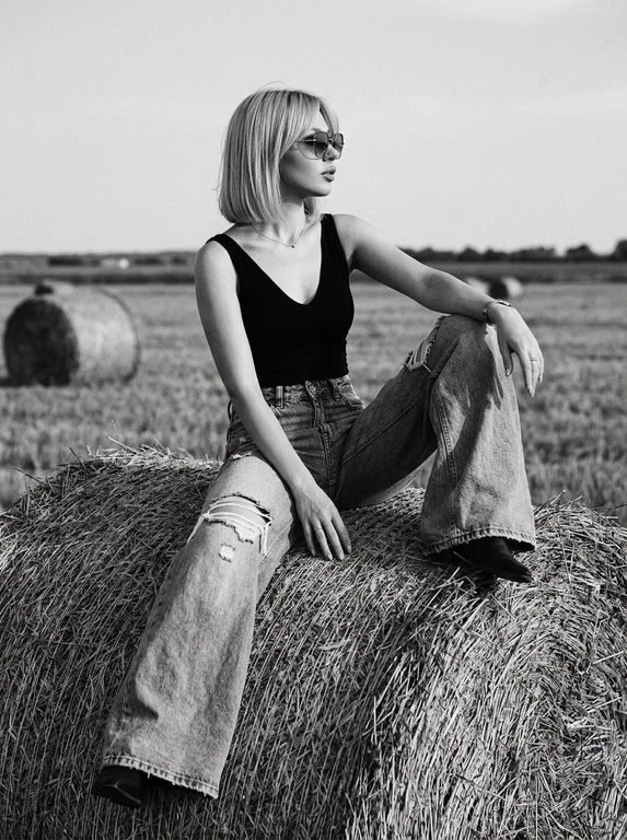 Чёрно-белая high-fashion editorial фотография в сельской местности, женщина с внешностью как на референсе, черты лица не изменять, композиция — более общий план (wide shot), камера на среднем расстоянии, фигура полностью в кадре с окружающим пространством, она сидит на большом тюке сена в поле, корпус слегка развернут, одна нога свободно опущена вниз, вторая согнута, создавая расслабленную, но выразительную позу, руки естественно лежат на колене и на поверхности тюка, взгляд направлен в сторону, профиль, выражение спокойное и уверенное, одежда: чёрный топ с V-образным вырезом и объёмные светлые широкие джинсы с потёртостями и рваными элементами, аксессуары: крупные солнцезащитные очки, минималистичные украшения, фон: открытое поле с несколькими тюками сена на разной глубине, создающими перспективу, линия горизонта ниже центра кадра, чтобы подчеркнуть небо, освещение: жёсткий естественный дневной свет с мягкими облаками, формирует выразительные тени и контраст, стиль: кинематографический, плёночное зерно, высокая детализация, реалистичная текстура кожи и ткани, редакционный fashion стиль, акцент на атмосфере и пространстве кадра


Какая будет прическа?: Волосы каре с челкой по бокам