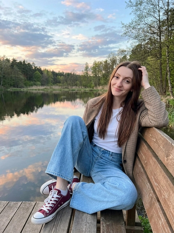 Портретная фотография на природе во время заката. Женщина сидит на деревянной скамейке со спинкой у берега спокойного водоема или реки. На заднем плане виден лесистый берег, а небо с облаками отражается в воде. Атмосфера кадра тихая, спокойная и умиротворенная. Цветовая гамма теплая и мягкая, с сочетанием голубых, розовых и оранжевых оттенков закатного неба.
На женщине белая футболка, голубые широкие оверсайз джинсы и бордовые кеды  Converse Chuck Taylor All Star OX с белыми шнурками. Поверх футболки накинут пиджак в клетку коричнево-бежевых тонов. Образ выглядит повседневно, естественно и расслабленно.
Поза непринужденная: женщина сидит на скамейке полулежа, слегка облокотившись на спинку. Одна рука поднята к голове, будто она поправляет волосы или просто касается виска в естественном жесте. Взгляд направлен прямо в камеру. Кадр передает ощущение уюта, тишины и вечернего покоя. Реалистичная фотография, мягкий естественный свет, живая атмосфера, натуральные цвета, спокойное lifestyle-настроение.