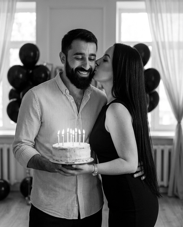 Create an ultra-photorealistic, cinematic black-and-white birthday portrait of a young romantic couple.
A man with, wearing a thin vertically striped button-up shirt, smiling naturally with his eyes closed. He holds a round white-frosted birthday cake with six lit candles at waist level.
A woman stands closely on his right, gently kissing his cheek with her eyes closed. wears an elegant fitted black sleeveless dress with a modest neckline and a delicate wristwatch. Her arm wraps naturally around his waist, while his arm embraces her.
Elegant indoor birthday setting with clusters of black balloons, large windows, sheer white curtains, and soft natural daylight. Intimate, romantic, spontaneous atmosphere.
Professional 85mm portrait photography, realistic skin pores, natural hair strands, authentic anatomy, soft cinematic lighting, rich monochrome contrast, subtle film grain, realistic candle flames, 8K photographic quality.
Negative: CGI, plastic skin, excessive smoothing, distorted faces, unnatural hands, extra fingers, artificial expressions, cartoon, beauty filters, unrealistic anatomy.

Доп. характеристики: Лица не меняй . У девушки длинные черные волосы до попы