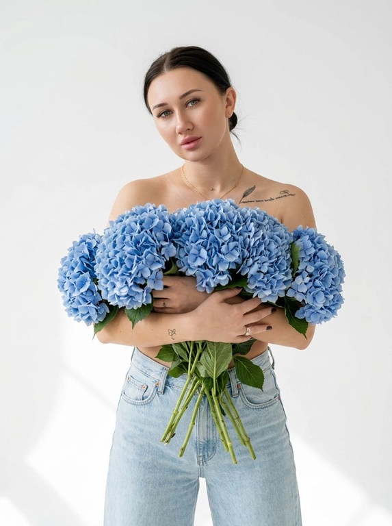 Женщина позирует в на белом фоне. Мягкие тени на стене и полу, ощущение воздуха и пространства. Макияж натуральный: ровный сияющий тон кожи, подчеркнутые ресницы, мягкие розовые губы. Атмосфера кадра — женственность, свежесть, легкость, весеннее настроение. Крупный план по грудь. На ней только светло-голубые широкие джинсы Levi’s с высокой посадкой. Взгляд в камеру, спокойное уверенное выражение лица. Голова немного наклонена вбок.

Какие будут цветы?: Она обнимает большой букет голубых гортензий, полностью прикрывая им грудь. Стебли естественно опущены вниз под весом цветов.