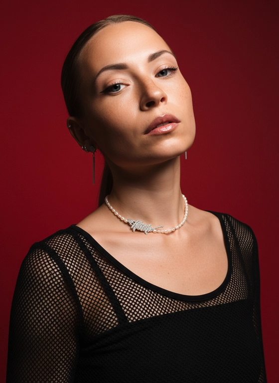 Создай портрет, используя те же черты лица (на основе загруженного фото), характеризующийся резким кинематографическим освещением и интенсивным контрастом. Снятый с немного нижнего ракурса, направленного вверх, что подчеркивает линию подбородка и шеи модели, этот портрет производит впечатление доминантности и скульптурной элегантности. Фон — глубокий, насыщенный малиново-красный, создающий яркий визуальный контраст со сияющей кожей модели и темной одеждой.