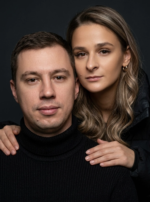 Гиперреалистичный студийный fashion-портрет молодой пары по загруженному референсу, 100% сходство лиц. Черты лица не изменять. Крупный план, кадр по плечи. Мужчина, спокойный уверенный взгляд прямо в камеру, естественное выражение лица, одет в черный трикотажный свитер с высоким воротом. Женщина стоит позади мужчины и нежно обнимает его, ее руки лежат на его плечах. Ее лицо расположено рядом с его лицом, она повернута слегка боком, голова немного наклонена вперед, взгляд направлен прямо в камеру. У женщины выразительные глаза с длинными густыми черными ресницами, естественный clean-glam макияж, натуральные брови, теплый оливковый оттенок кожи, небольшие серьги-кольца. Композиция плотная и интимная, их лица расположены очень близко друг к другу, современный модный портрет пары. Женщина одета в черный корсет. Кожа максимально натуральная: живая текстура кожи, видимые микропоры, subtle skin glow, естественный мягкий блеск кожи, natural skin microtexture, skin translucency, micro skin details, легкие естественные несовершенства кожи, без ретуши и без эффекта пластика. Освещение: мягкий студийный beauty lighting, рассеянный ключевой свет спереди и немного сбоку, мягкие глубокие тени для объема лица, деликатные световые блики на коже. Фон: глубокий темный студийный фон, темно-графитовый или почти черный, создающий контраст с кожей и лицами. Стиль: luxury fashion editorial photography, high fashion portrait, ultra photorealistic, 85mm portrait lens, shallow depth of field, creamy bokeh, cinematic soft lighting, RAW photo, extremely detailed, fine hair strands, natural skin pores, professional photography, photorealism, 8k, high dynamic range.

Доп. характеристики: Добавить легкий макияж  и сделать легкие волнистые локоны губы не увеличивать