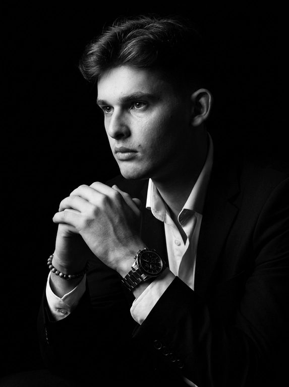A hyper-realistic 8K cinematic portrait of a uploaded person,
captured in a dramatic Chiaroscuro style. The subject is seated at a
three-quarter angle, leaning slightly forward with a relaxed yet
commanding posture. His face is turned away from the lens, with
one side of his face clearly illuminated while the other is
completely lost in deep, velvety black shadows. His expression is
contemplative, not looking at the camera. The pose is precise: his
hands are clasped together near his chest; one hand is held
straight with fingers slightly tilted, while the other hand is
positioned so only the fingers are visible, resting over the first. He
wears a luxury black chronograph watch with a detailed metal link
bracelet, and a beaded bracelet. His attire is a sharp
black suit jacket over a white dress shirt with the top buttons
open, showing refined fabric textures. The background is a solid,
seamless black. The lighting is sharp and directional, highlighting
the intricate details of his hair,
the moisture in his eyes, and the glint off the watch face.
Cinematic black-and-white color grade with rich contrast and
smooth, natural skin transitions. Shot on 85mm lens, f/1.8, high-
resolution commercial photography