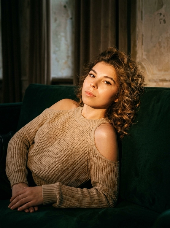Абсолютное сохранение черт лица модели, внешность 1:1, не искажать и не менять лицо.
Общая стилистика:
Глубокий, атмосферный, интимный fashion editorial портрет с кинематографическим качеством. Изображение должно быть "moody" (настроенческим), с высоким контрастом и ощущением пленочной фотографии.
Поза и Ракурс:
Женщина в расслабленной позе полулёжа откинулась назад на винтажном бархатном диване глубокого цвета. Корпус развернут в 3/4, одна рука служит опорой за спиной, ноги согнуты в коленях и перекрещены. Ракурс камеры чуть ниже уровня глаз, создавая ощущение подглядывания. Кадрирование по бедра (3/4 роста), основной акцент на ключицы, плечи и зону декольте.
Внешность и Одежда (Без указания цвета волос):
Макияж: Акцент на длинные, разделенные черные ресницы. Мягкий скульптурирующий контуринг лица, естественный румянец. Заметный влажный хайлайт на ключицах и плечах.
Освещение и Интерьер:
Свет: Драматичное, теплое боковое освещение (как от окна на закате) служит рисующим светом, создавая глубокие тени и подчеркивая объем. Тонкий, слабый контровой свет (rim light) едва очерчивает силуэт плеч и прически, отделяя модель от фона.
Окружение: Комната в полумраке. На фоне угадываются тяжелые фактурные портьеры, старая штукатурка на стене или деревянные панели. Атмосфера будуара или старой квартиры.
Технические и Художественные Детали:
Эстетика пленочного кадра, снятого на объектив 35mm или 50mm с открытой диафрагмой (f/2.8), обеспечивающей мягкое, художественное боке на заднем плане. Изображение слегка недоэкспонировано (low key) для насыщенности теней. Цветокоррекция в тёплых, приглушённых бежево-коричневых и охристых тонах с пониженной насыщенностью холодных оттенков (зеленого и синего). Высокая детализация текстуры кожи, ткани денима и сатина, подчёркнутая легкой, естественной пленочной зернистостью и мягкой виньеткой по краям кадра.

Какая будет прическа?: Волосы уложены в объёмные локоны

Какая будет одежда?: Вязанные свитер бежевого цвета с открытыми плечами и рукавами.
