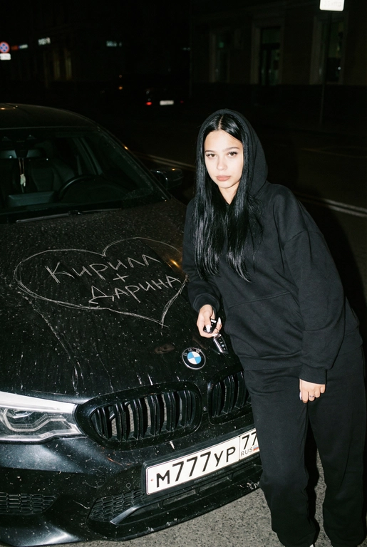 Строго сохранить лицо с прикреплённой референс-фотографии, на 100% без искажений. Человек одет полностью в чёрный, монохромный оверсайз спортивный костюм: свободная худи на молнии и широкие расслабленные штаны, без логотипов, текста и брендинга. Акцент на реальной текстуре кожи и детализированной текстуре ткани.
Сырой кинематографичный ночной кадр. Человек опирается на капот автомобиля, средний план, лёгкий верхний ракурс. В одной руке игриво держит ключи от машины. Номерной знак виден: m777yp 77.
Строго по центру капота, покрывая всю поверхность капота, глубоко и грубо процарапаны (не мелом) царапины: большое сердце с текстом «» внутри.
Жёсткая прямая вспышка, сильные тени, высокий контраст, видимая пыль и микро-царапины на краске. Тёмный размытый городской фон.
Объектив 35mm, f/2.8, грубая уличная эстетика.

Надпись на капоте: Кирилл +Дарина

Марка автомобиля: Опирается на чёрный BMW M6 F90