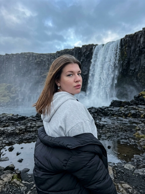 Максимально реалистичная lifestyle-фотография женщины, снятая на iPhone. Натуральное RAW-качество, слегка затемнённая холодная экспозиция, лёгкая естественная зернистость, реалистичная мобильная оптика и естественный динамический диапазон.

Женщина стоит спиной к камере в естественном полуобороте. Корпус развёрнут от объектива, голова мягко повернута назад через плечо так, чтобы лицо читалось примерно в три четверти. Взгляд направлен в сторону камеры, спокойный, уверенный и немного загадочный. Поза выглядит непринуждённо, будто это случайный кадр во время путешествия, без ощущения постановочной fashion-позы.

Волосы слегка двигаются от холодного ветра.

На женщине очень объёмное светло-серое худи oversize из мягкой плотной ткани со свободным силуэтом. Поверх плеч небрежно спущена большая чёрная матовая пуховая куртка, очень объёмная и мягкая, свободно лежащая на плечах и руках. Образ расслабленный, тёплый и контрастный на фоне холодного вулканического пейзажа. 

Макияж выразительный, но реалистичный: естественная кожа с видимыми порами, лёгкой неоднородностью тона и натуральными переходами света и тени. Мягкое естественное сияние только на выступающих участках лица. Скулы слегка подчёркнуты, без жёсткого контуринга. Тонкие вытянутые чёрные стрелки, выразительные, но реалистичные ресницы. Брови аккуратно уложены. Губы естественной формы, пудрово-розового оттенка, с лёгким влажным блеском. Без бьюти-фильтра, фарфоровой кожи и глянцевого AI-макияжа.

Фон — огромный тёмный вулканический ландшафт из мокрых чёрных и графитовых матовых камней. Поверхность неровная, с естественными трещинами и небольшими лужами. В центре заднего плана находится мощный высокий водопад, низвергающийся между массивными тёмными скалами. У основания скал висит густой холодный туман и водяная дымка.

Небо пасмурное и слегка затемнённое, с тяжёлыми серо-синими облаками. Холодная приглушённая палитра: графитовый, серый, сине-стальной и чёрный. Без зелени, яркой растительности и насыщенных цветов.

Свет полностью естественный, пасмурный и рассеянный. Лицо освещено мягко и неравномерно, без студийного бьюти-света. Сохраняются небольшие естественные тени под глазами, возле носа и скул. Фон слегка недоэкспонирован, женщина немного лучше читается за счёт естественного рассеянного света.

Фотореализм, натуральная текстура кожи, отдельные волоски, реалистичные фактуры материалов и мокрых камней, лёгкий цифровой шум, приглушённые цвета. Ощущение настоящей фотографии из путешествия, снятой человеком на телефон. Без CGI, HDR-эффекта, пластиковой кожи, excessive skin smoothing, beauty filter, AI-gloss и чрезмерной резкости.

Доп. характеристики: Волосы прямые ухоженные