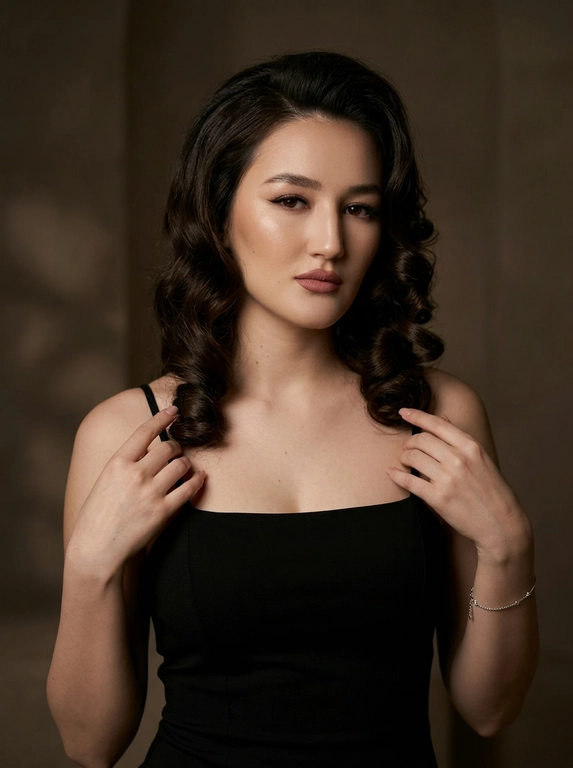 Ultra-realistic cinematic fashion portrait, moody editorial style. Elegant, refined, sophisticated atmosphere. Waist-up shot. A woman in soft low light, both hands raised near the collarbone, fingertips lightly touching the thin straps of her top, elbows relaxed and lowered, shoulders soft. Head slightly turned to the side, thoughtful gaze directed away, calm and confident expression. Long dark hair falling over one shoulder. Evening glam makeup: defined winged eyeliner, full lashes, soft face contouring, dusty-rose matte lipstick. Fitted black sleeveless top with thin straps and a square neckline, matte fabric; delicate silver bracelet on the wrist. Minimalist warm dark-toned interior, plain softly textured background fading into deep shadow, subtle warm light on the wall behind. Low-key lighting, soft warm side light emphasizing the collarbones, shoulder line and cheekbones. Deep cinematic shadows for volume and dramatic mood. 85mm f/1.4, shallow depth of field, focus on the eyes and face, soft background blur. Natural realistic skin texture, visible pores, no plastic retouching.

Какая будет прическа?: Уложенные в крупные классические локоны с заметным объемом у корней. Прическа выглядит аккуратной и глянцевой.