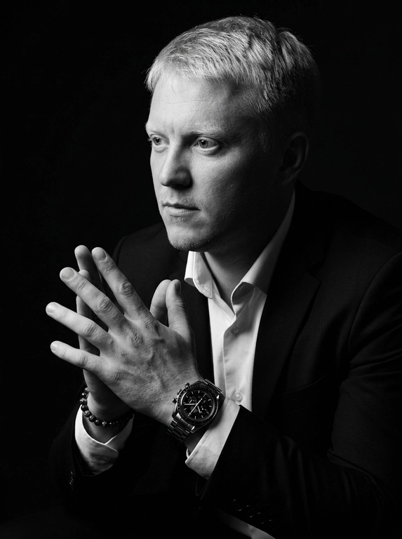 A hyper-realistic 8K cinematic portrait of a uploaded person,
captured in a dramatic Chiaroscuro style. The subject is seated at a
three-quarter angle, leaning slightly forward with a relaxed yet
commanding posture. His face is turned away from the lens, with
one side of his face clearly illuminated while the other is
completely lost in deep, velvety black shadows. His expression is
contemplative, not looking at the camera. The pose is precise: his
hands are clasped together near his chest; one hand is held
straight with fingers slightly tilted, while the other hand is
positioned so only the fingers are visible, resting over the first. He
wears a luxury black chronograph watch with a detailed metal link
bracelet, and a beaded bracelet. His attire is a sharp
black suit jacket over a white dress shirt with the top buttons
open, showing refined fabric textures. The background is a solid,
seamless black. The lighting is sharp and directional, highlighting
the intricate details of his hair,
the moisture in his eyes, and the glint off the watch face.
Cinematic black-and-white color grade with rich contrast and
smooth, natural skin transitions. Shot on 85mm lens, f/1.8, high-
resolution commercial photography