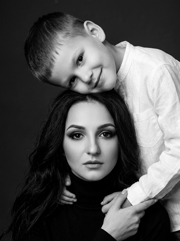 Родитель и ребёнок с внешностью как на приложенных фото, художественная семейная студийная фотосессия. Композиция — вертикальный портрет, крупный план лиц. Родитель внизу смотрит прямо в камеру, лицо фронтально. Ребёнок сверху, голова повернута набок, щека лежит на голове родителя, смотрит в камеру с нежной улыбкой. Рука ребёнка обнимает родителя за шею, видна в кадре, родитель держит руку ребёнка своей рукой.
Одежда: ребёнок в белой льняной рубашке, родитель в чёрной водолазке. 
Фон — тёмно-серый почти чёрный однотонный студийный, создаёт контраст. Освещение — мягкое боковое студийное, создаёт объём и глубину.
Чёрно-белая художественная фотография с выразительными контрастами. Атмосфера нежная, интимная, спокойная.

Доп. характеристики: Элегантный чёрно-белый крупный план студийного портрета, красивая молодая мама с длинными тёмными волосами и выразительным макияжем глаз лежит или сидит, её 8–10-летний сын с короткой стрижкой опирается сверху, щека к щеке или лоб ко лбу, оба спокойно и уверенно смотрят прямо в камеру с любящими выражениями