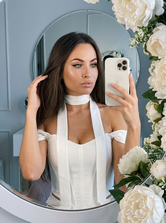 Максимально реалистичное селфи в зеркале в эстетике luxury soft glam. Модель стоит перед круглым зеркалом с изящной рамой, украшенной белыми живыми пионами. Цветы обрамляют зеркало нежно и дорого, создавая ощущение утончённого, премиального кадра. Композиция чистая и визуально собранная: модель находится в центре отражения, кадр выглядит эстетично, дорого и современно.

Поза естественная и уверенная: одной рукой модель держит современный смартфон, второй рукой слегка касается волос, создавая живой и непринуждённый жест. Селфи выглядит не постановочным, а как красивый спонтанный кадр из дорогой life-style съёмки.

Макияж — выразительный soft glam: чёткие чёрные стрелки, длинные пышные ресницы с лисьим эффектом, внутренние уголки глаз подсвечены сияющими тенями. Кожа ровная, ухоженная, с мягким влажным сиянием. Губы подчёркнуты карандашом и покрыты полупрозрачным глянцевым блеском, создающим аккуратный объём.

На модели ее образ выглядит женственно, современно и дорого. 

Смартфон современный, в минималистичном белом матовом чехле. Если нужен более дорогой вайб, без лишнего декора и ярких деталей.

Освещение холодное, мягкое и чистое, как в дорогой beauty-съёмке или premium dressing room. Свет красиво подчеркивает сияние кожи, текстуру ткани, свежесть пионов и блеск украшения. Атмосфера — роскошная, женственная, ухоженная, без дешёвого глянца.

Фотореализм максимальный: высокая детализация кожи, ткани, цветов, аксессуаров и отражения в зеркале. Лёгкий цифровой шум, мягкая резкость, деликатная зернистость, естественная глубина кадра, высокая контрастность, эффект живого фото, без CGI и без искусственного AI-эффекта.

Какая будет прическ?: Не менять мою прическу

Какой будет образ на фото? : Белый топ-корсет с открытыми плечами. Его главная особенность — длинная лента (или шарф), выполненная из той же ткани, что и топ. Лента плотно охватывает шею, создавая эффект шарфа, прямые и свободные концы ленты спускаются вниз они проходят вдоль передней части топа, свободно свисая.  Образ элегантный.  Акцент на линии шеи и вертикальных линиях торса.