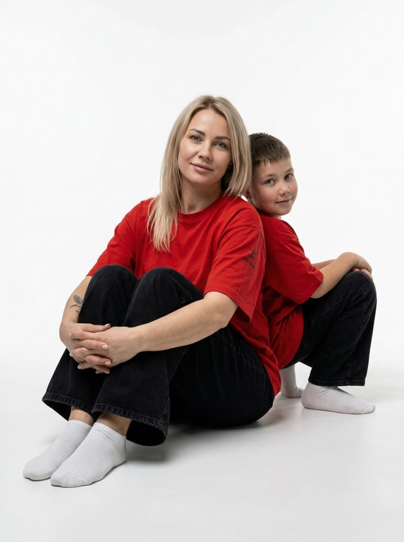 Гиперреалистичная фотография. Качество 8К. Используя портретное сходство с людьми с фотографий, создай фотографию: женщина в красной оверсайз свободной футболке и свободных черных джинсах, белых носках. Ребенок одет в красную оверсайз футболку, черные свободные джинсы и белые носочки. Женщина сидит на полу, согнув колени, смотрит прямо в кадр с еле заметной улыбкой. Сзади женщины в такой же позе сидит ребенок облокотившись спиной к спине женщины, с легкой улыбкой. Темный стиль модного редакционного журнала, глубокие тени и драматическое студийное освещение, белый фон, элегантная и мощная композиция, ультрареалистичный, высокая детализация, художественный портрет.