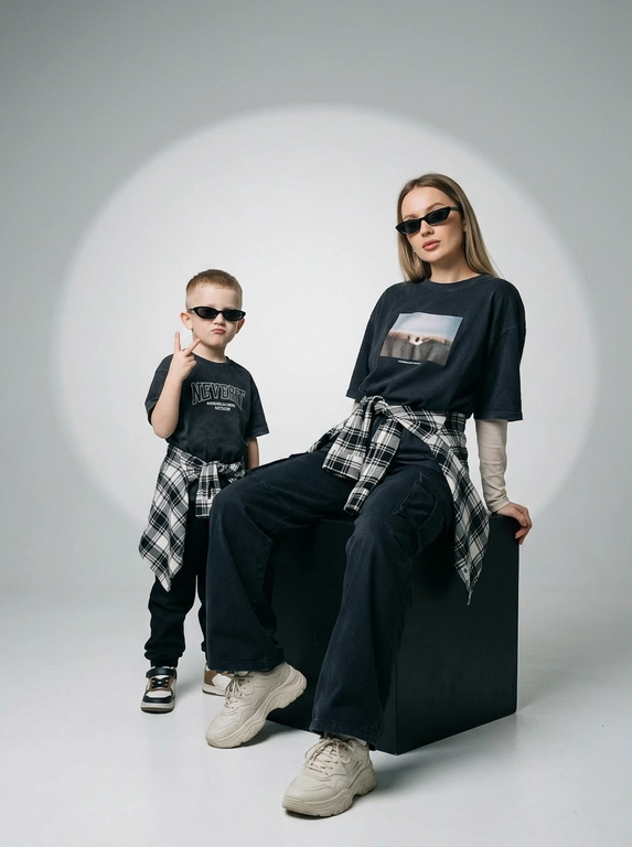 Photorealistic studio portrait photography in minimalistic fashion/editorial aesthetics, static camera at chest level of a seated person, light frontal angle without noticeable perspective deformation; composition with two characters: an model sits on a large matte black cube, body slightly turned towards the camera, knees bent, feet on the floor, the arms are lowered and relaxed in front of the knees, the fingers are neatly extended; the child stands on the same cube behind and to the left, at full height, one hand raised with a "V" gesture, the other lowered, pose confident and playful; both wearing narrow dark sunglasses, gaze directed at the camera; shooting plan — full height for both with a small supply of air above and below, center the center of the frame is shifted to the right to a sitting figure, a standing child balances on the left; the lens is normal/short portrait 50-70 mm, without width; aperture is approximately f/5.6–f/8, moderate depth of field, both characters are sharp, the background is slightly soft; the studio lighting is soft, with one key source on the front left at a level just above the head (softbox), giving smooth transitions and a pronounced but soft shadow against the background on the right; light filling light from the front reduces contrast, shadows are readable; the background is a smooth, light, seamless cyclorama/wall with a noticeable large oval light spot (gradient) in the center, there is a darker vignetting around, a clean minimalistic studio without props; the mood is calm, stylish, slightly retro, with an emphasis on street fashion and family vibe; the expression on the face of the seated model is a soft smile, relaxation; the person standing has a confident neutral facial expression; clothes: the seated model has a dark oversized T-shirt with a large rectangular print on the chest, under it a light longsleeve with long sleeves, wide dark cargo trousers/joggers with pleats at the knees and bottom, a plaid shirt/flannel tied at the waist with a knot, massive light sneakers with a pronounced sole and mesh texture; the child has a dark T-shirt with a large inscription on the chest, dark trousers, a plaid shirt tied at the waist, sneakers in two colors; fabrics with a realistic cotton and flannel texture, noticeable fine folds, natural creases on the knees and in the waist area, matte surfaces without gloss; micro-details: natural skin texture without plastic, neat glare on the lenses of glasses, a clear contour of the shadow from the raised hand on the background, clean floor and soft drop shadow from the cube; high detail, natural sharpness, light grain like a film shot, moderate contrast, soft matte/film look, no HDR, no overexposure and no unnecessary objects.

Какого цвета будут рубашки на поясе?: Чёрно-белый.