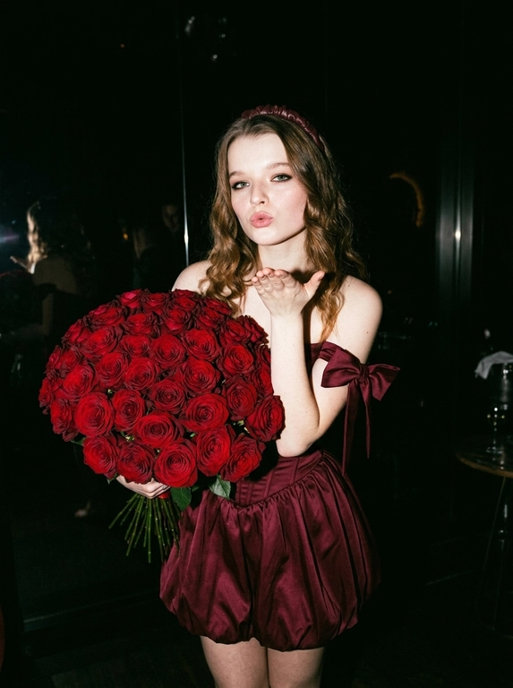 Сохранить внешность на 100%. Черты лица не изменять.
Ultra-realistic vertical 9:16 photo, Y2K luxury nightlife, paparazzi flash.

Полная темнота.
Фон почти полностью чёрный — ресторан не читается, только намёки: слабые отражения, едва заметные силуэты, глянец поверхностей. Всё пространство «съедено» тьмой.

Единственный источник света — жёсткая вспышка камеры, бьёт прямо в лицо и фигуру, создаёт резкий контраст, глубокие тени, эффект ночного paparazzi-кадра начала 2000-х.

Женщина в платье:
— глубокий винно-бордовый / dark cherry цвет
— корсетное мини без бретелей
— жёсткий sculptural corset
— короткая объёмная bubble-юбка
— off-shoulder банты
— плотная дорогая couture-ткань
— силуэт 1:1, без изменений

В руках — огромный, сверхпышный букет цветов, почти перекрывает корпус:
— тяжёлый, статусный,
— максимально плотный,
— luxury florist look.

Действие:
— она посылает воздушный поцелуй в камеру,
— взгляд дерзкий, холодный, уверенный,
— без улыбки.

Эффект:
— вспышка «вырывает» её из темноты,
— кожа и ткань ловят свет,
— фон проваливается в чёрный.

Стиль:
Y2K rich bitch, flash-only lighting, dark luxury, paparazzi moment, ultra-realistic, expensive, iconic, no softness, no romance, pure power.

Какие будут цветы: В руках у нее букет тёмно-красных роз.