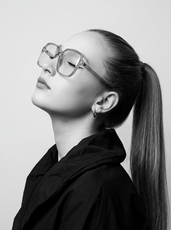 Чёрно-белый минималистичный фотореалистичный портрет, студийный мягкий свет спереди‑сверху, нейтральная светлая однотонная стена как фон. Девушка в профиль: голова запрокинута назад, подбородок поднят, глаза закрыты — расслабленная fashion-поза с акцентом на линии шеи и контуры лица. Обязательно точно сохранить черты лица с референс-фото (без искажений); крупные квадратные очки в массивной полу-прозрачной оправе с естественными бликами. Детали лица: длинные ресницы и чёткие стрелки, выразительные губы, естественная текстура кожи, аккуратный ретуш. Одежда: тёмный объёмный верх с выразительным воротником, маленькая серьга-кольцо. Стиль: высокая графичность и контраст, чистые линии, лёгкая драматичность, максимальная детализация и фотореализм, аккуратные светотеневые переходы, подчёркнутые контуры.

Какая будет прическа?: Гладкие волосы, высокий длинный густой хвост.