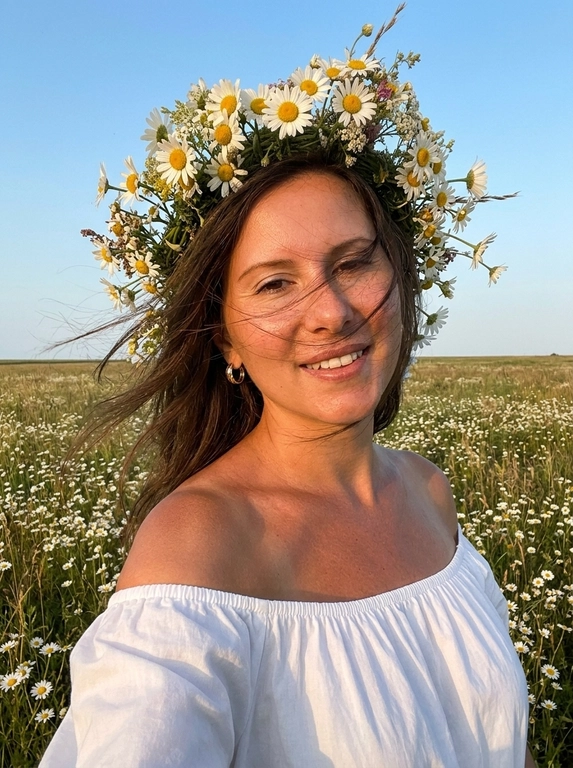 Женщина делает селфи. На ней белое легкое платье с открытыми плечами, лёгкое хлопковое  платье. Кожа загорелая. У нее на голове объёмные цветочные венки из мелких полевых ромашек . Венки должны быть пышные, натуральные, свежие, как летний flower crown. Цветы расположены вокруг головы, без искусственного вида.
Её волосы развиваются от ветра слегка падая на лицо.
Фон: летнее поле с ромашками. Открытое небо без облаков слегка голубое.
Не размывай фон. Голова слегка наклонена к плечу.
Рук в кадре нет.
Горизонт прямой , волосы сильно развиваются от ветра. Солнечная погода в золотой час.