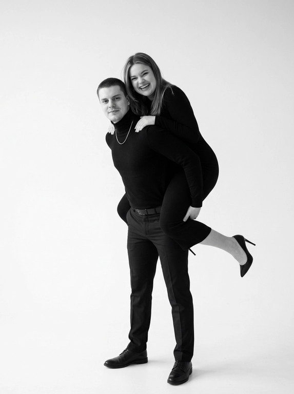 Фотография в студийном стиле, с минималистичным белым фоном. Два человека: мужчина и женщина, запечатлены в динамичной позе. Цветовая гамма – монохромная, с преобладанием черного и белого, создающая контрастность и акцент на форме.

Мужчина одет в костюм, состоящий из водолазки и классических брюк, на ногах черные туфли. Он стоит прямо, немного согнув ноги в коленях. Руками он держит женщину, перекинутую через его спину.

Женщина одета в облегающее платье с длинными рукавами. На ногах черные туфли-лодочки на высоком каблуке. Одна нога вытянута вперед. Ее поза выражает легкость и игривость. Она смотрит в камеру с улыбкой.

Атмосфера фотографии передает непринужденность и веселье. Освещение ровное, без резких теней, что подчеркивает гладкость фона и чистоту линий.