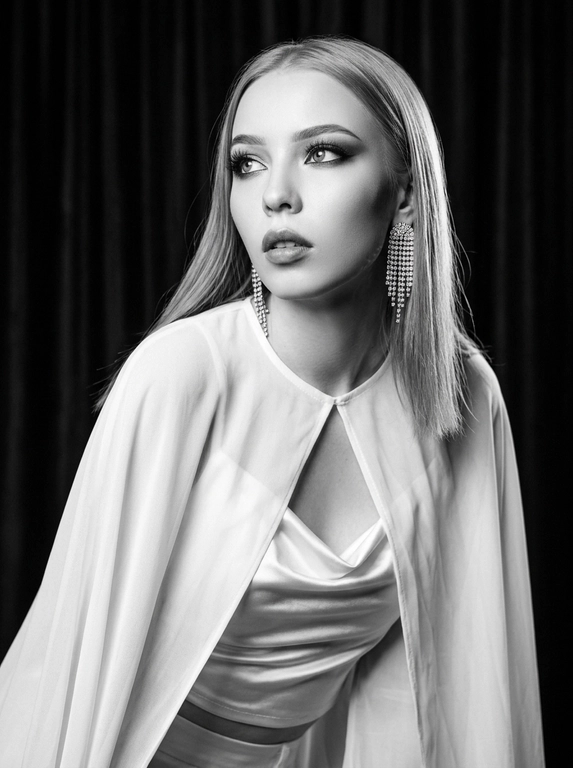 Фотореалистичный контрастный чёрно-белый glamour/editorial портрет в стиле Old Hollywood, вертикальный кадр по грудь. Женщина в 3/4, корпус чуть наклонён вперёд; голова повернута влево, взгляд вверх/в сторону (не в камеру), губы чуть приоткрыты, выражение драматичное и кинематографичное. Образ: белая накидка из шифона с длинными струящимися рукавами, под накидкой драпированный атласный белый топ; на ушах крупные серьги со стразами/каскадом. Макияж: классический вечерний glam (в ч/б читается как контраст) — чёткие брови, мягкие smoky тени, тонкая стрелка, длинные ресницы, губы с чётким контуром и сатиновым финишем. Фон: тёмный студийный с мягким рисунком/драпировкой (как бархатные шторы), размытый. Свет: жёсткий key слева спереди (beauty dish) + контровой/rim справа для отделения волос, глубокие тени, яркие блики на скулах и волосах, богатый тональный диапазон (глубокие чёрные и сливочные белые). Камера: 85mm, f/2.0–2.8, ISO 100–200, 1/160, резкость на глазах и губах, фон в мягком боке, лёгкое плёночное зерно, без “пластика”.

Какая будет укладка? : Длинные светло-русые волосы 

Доп. характеристики: Цвет глаз зеленый, волосы прямые светло-русые длинные