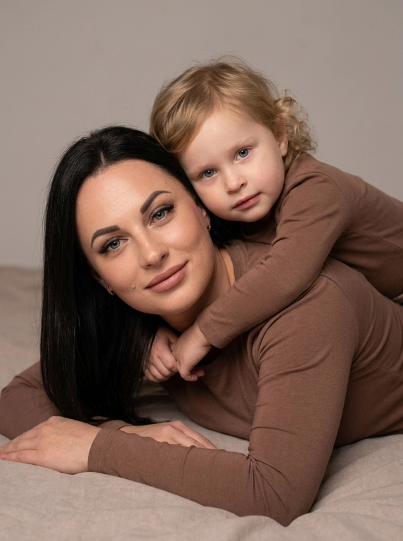 Создай студийный портрет родителя и ребёнка, максимально точно сохранив черты лица и выражения с референсных изображений. Не изменяй и не искажай лица, воспроизведи их с фотографической точностью.

Композиция:

Крупный план, ракурс на уровне глаз.

Родитель и ребёнок лежат на животе, расположены боком к камере.

Ребёнок лежит на спине родителя и мягко обнимает его за плечи.

Оба смотрят прямо в камеру, создавая тёплый и близкий зрительный контакт.

На родителе и ребёнке одинаковые коричневые лонгсливы без принтов и декора.

Естественный внешний вид, ровный тон кожи.

Мягкое студийное освещение, спокойная семейная атмосфера, натуральная текстура кожи, высокая детализация, максимальный фотореализм. Без ламп в кадре и прочего декора.

Формат 9:16.

Качество 8K.