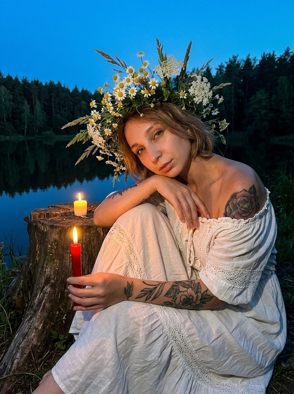 Ultra-realistic Ivan Kupala night atmosphere near a calm lake at twilight. Woman sitting beside an old tree stump near the water, body slightly leaning forward in a dreamy relaxed pose, one arm resting naturally over the knee, the other delicately holding a thin burning red candle beside another candle on the stump. White off-shoulder vintage peasant dress with lace details and soft fabric folds. Hair styling only: soft natural styling with loose face-framing strands. Large realistic wildflower crown made of chamomile flowers, meadow grass and white blossoms. Dark forest and calm lake behind, warm candlelight illuminating the face and hands, deep blue twilight shadows, mystical summer night mood. Shot on iPhone night mode, realistic low-light grain, lowered exposure, no flash, no blur, no bokeh, everything sharp and in focus.