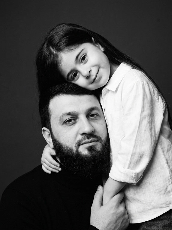 Родитель и ребёнок с внешностью как на приложенных фото, художественная семейная студийная фотосессия. Композиция — вертикальный портрет, крупный план лиц. Родитель внизу смотрит прямо в камеру, лицо фронтально. Ребёнок сверху, голова повернута набок, щека лежит на голове родителя, смотрит в камеру с нежной улыбкой. Рука ребёнка обнимает родителя за шею, видна в кадре, родитель держит руку ребёнка своей рукой.
Одежда: ребёнок в белой льняной рубашке, родитель в чёрной водолазке. 
Фон — тёмно-серый почти чёрный однотонный студийный, создаёт контраст. Освещение — мягкое боковое студийное, создаёт объём и глубину.
Чёрно-белая художественная фотография с выразительными контрастами. Атмосфера нежная, интимная, спокойная.