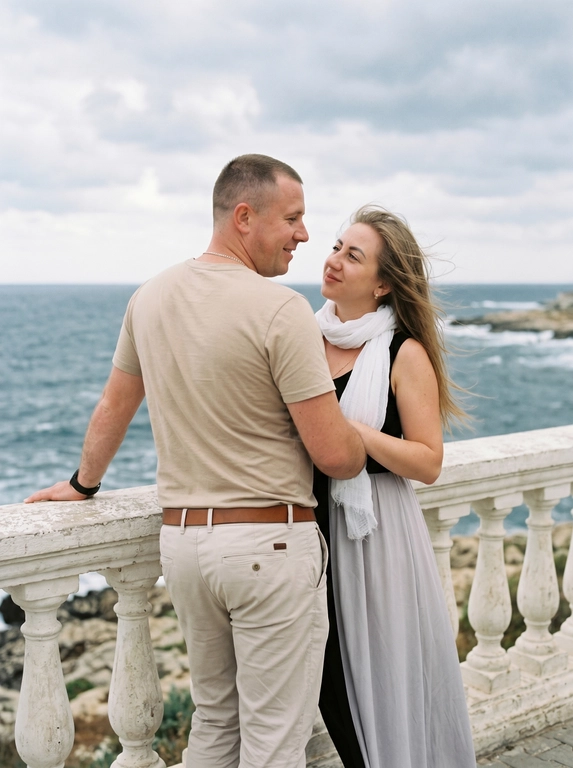 Пара стоит у белого каменного балюстрадного ограждения с видом на море. Мужчина слева, спиной к камере, корпус в 3/4 вправо, левая рука опирается на перила, голова повернута вправо к женщине, смотрит на неё с нежностью. Женщина справа, корпус в 3/4 влево, стоит очень близко к мужчине, смотрит ему в лицо снизу вверх. Одежда: мужчина — светлая бежевая футболка с коротким рукавом, светлые брюки, коричневый ремень; женщина — длинное платье с чёрным облегающим верхом без рукавов и светлой струящейся юбкой до пола, белый тонкий шарф на шее. Волосы слегка развеваются. Фон: море, пасмурное небо. Мягкий естественный свет, 50mm, лёгкое зерно.