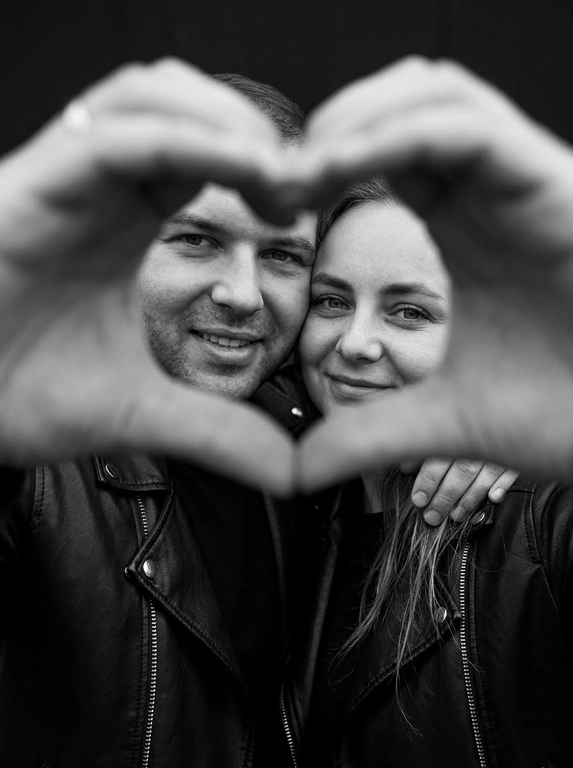 Ultra-realistic editorial photography of a smiling couple in a romantic and cinematic composition. The couple is centered in the background of the image, looking directly at the camera with a spontaneous, happy, and natural expression. In the foreground, extremely close to the lens and slightly out of focus, the couple’s hands form a heart shape perfectly framing their faces.

The composition creates strong visual depth, with sharp focus on the eyes and faces inside the heart formed by the hands. The hands occupy a large portion of the foreground, artistically blurred around the edges, creating a sophisticated cinematic effect.

Intimate, emotional, and elegant aesthetic inspired by modern perfume campaigns and premium lifestyle editorials. Soft studio lighting with delicate contrast and natural shadows. Minimalistic dark blurred background.

The couple wears minimalistic black leather jackets and sophisticated dark clothing. Genuine expressions, strong emotional connection, contemporary romantic atmosphere. Hyper-realistic skin texture, visible pores, detailed hair strands, authentic human appearance.

Black and white photography, rich tones, deep blacks, and soft highlights. 85mm lens, shallow depth of field, cinematic portrait photography, luxury editorial style, ultra detailed, hyper realistic, 8K.