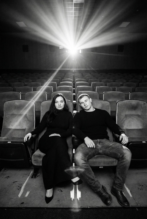  Кинематографичный чёрно-белый портрет пары в пустом кинозале.
Женщина и мужчина сидят рядом в мягких креслах кинотеатра.
кадр с глубоким пространством и пустыми рядами кресел позади них. Женщина черном длинном обтягивающем платье, поза женственная и спокойная, одна рука на подлокотнике.
Мужчина в черном свитере и в джинсах, сидит расслабленно.
Над ними яркий луч проектора, и выразительные блики.