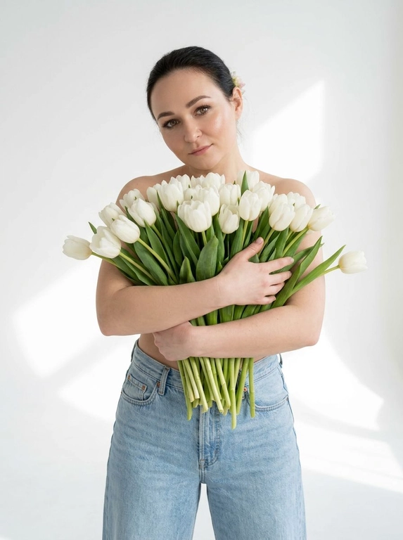 Женщина позирует в на белом фоне. Мягкие тени на стене и полу, ощущение воздуха и пространства. Макияж натуральный: ровный сияющий тон кожи, подчеркнутые ресницы, мягкие розовые губы. Атмосфера кадра — женственность, свежесть, легкость, весеннее настроение. Крупный план по грудь. На ней только светло-голубые широкие джинсы Levi’s с высокой посадкой. Взгляд в камеру, спокойное уверенное выражение лица. Голова немного наклонена вбок.

Какие будут цветы?: Она обнимает большой букет белых тюльпанов, полностью прикрывая им грудь. Стебли естественно опущены вниз под весом цветов.