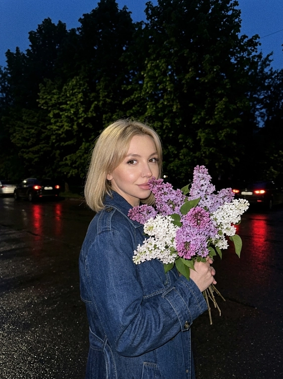 Очень крупный план, на этом фото женщина стоит в анфас к камере, обеими руками она прижимает к себе огромную пышную охапку сирени белой и фиолетовой, её голова слегка наклонена к цветам создавая ощущение что она вдыхает их аромат смотря в камеру, макияж: глаза подчеркнуты чёрными стрелками и длинными ресницами, на губах блеск, фон: действие происходит на улице в глубоких сумерках или ночью на заднем плане видны тёмные массивные силуэты деревьев с густой майской листвой внизу выкрашенные в белый цвет земля под ногами выглядит как тёмный асфальт после дождя на заднем плане едва заметные красные блики возможно огни далёких машин, ракурс: съёмка ведётся со стороны чуть сбоку на уровне глаз, освещение: слабое ночное освещение, фотореализм высокая детализация фото снято на камеру iPhone 17 Pro эффект живого кадра лёгкий цифровой шум зернистость мягкая резкость высокая детализация, без вспышки.

Какая будет одежда?: Темно-синий джинсовый тренч