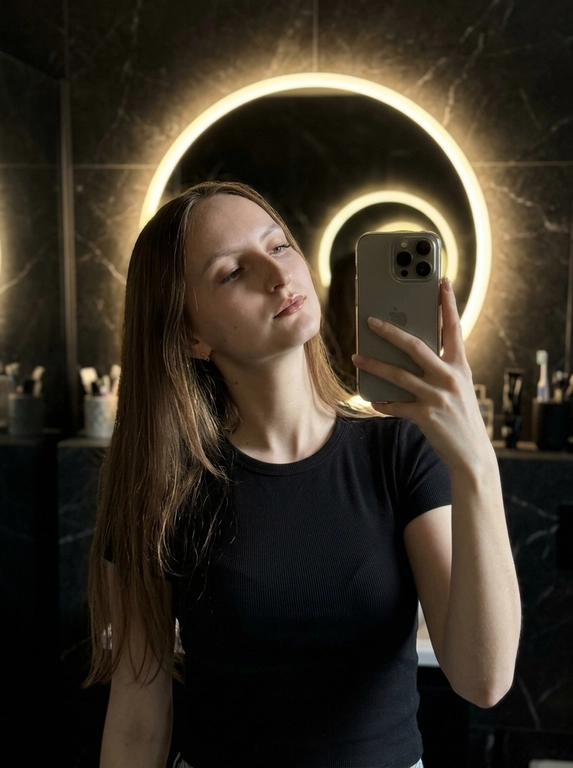 Снято на iPhone.
Женщина делает селфи в зеркало, в интерьере ванной комнаты.
Она слегка наклонила голову в сторону и назад. Взгляд в бок.
Освещение и тональность: тёплое, рассеянное освещение от круглого зеркала с подсветкой создаёт мягкое свечение по контуру головы и плеч, при этом спереди видно мягкие тени, подчёркивающие рельеф ключиц и декольте.
Интерьер и фон: стены облицованы темным мраморным кафелем.
 Фотография темная. Свет только от зеркала сзади. Не размывай фон. Без текста и водяных знаков. Без лишних рук, без ИИ глянца.

Какая будет одежда?: Чёрная приталенная футболка в рубчик

Какая будет укладка?: Не менять мою прическу