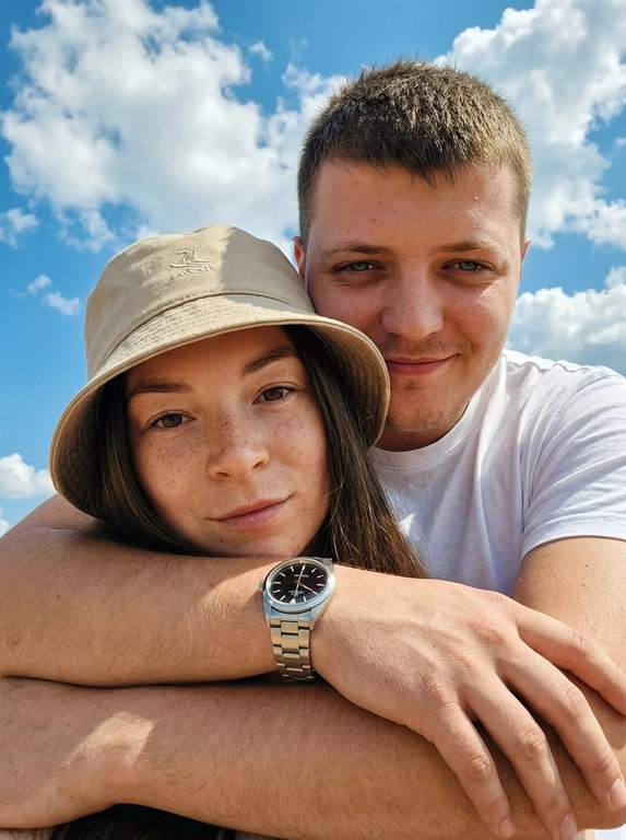 Кинематографичный кадр крупным планом (selfie style), пара в объятиях (классическое "обнимашки сзади") на фоне ярко-голубого неба с облаками.  Женщина на ней бежевая панама с брендовой надписью. Её кожа выглядит сияющей, подсвеченной мягким естественным солнечным светом. Выражение лица спокойное, глубокий взгляд, направленный прямо в камеру. Мужчина стоит строго позади девушки и обнимает её сзади, одна его рука обнимает её за плечи/шею спереди — предплечье лежит поперёк её ключиц, кисть свободно свисает с её плеча, на запястье классические серебряные часы с черным циферблатом. Мужчина в белой футболке, выражение лица доброжелательное, его взгляд прямо в объектив. Ракурс сверху, камера сфокусирована на лице девушки. Высокий контраст, насыщенные цвета, мягкая прорисовка деталей кожи. Эффект пленочной фотографии, легкое зерно, атмосфера летнего дня, эстетика социальных сетей. Телефона не видно в кадре.

Доп. характеристики: Без макияжа точь в точь как на фотографии