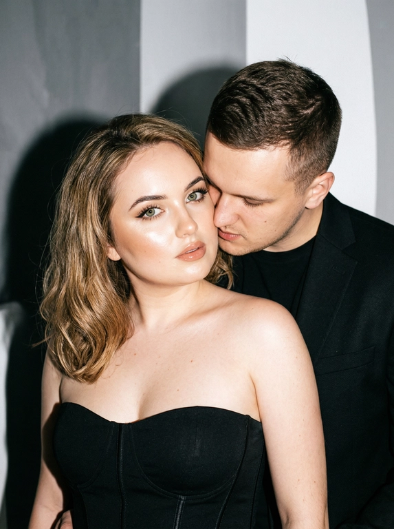 Не меняя черты лица, студийная fashion-съемка пары в минималистичном интерьере с серо-белым фоном, атмосфера гламура и интимной близости, ракурс — крупный портрет по грудь с легким поворотом корпуса, женщина на переднем плане, мужчина позади склоняется к её плечу, поза — она слегка отвернута от камеры с прямым взглядом, он касается лицом её шеи/плеча, одежда — на женщине черный корсет/бандо с открытыми плечами, на мужчине классический черный пиджак и рубашка, макияж — glam (ровный сияющий тон, сильный хайлайтер, четкий контуринг, стрелки, густые ресницы, глянцевые губы), свет — жесткая фронтальная вспышка + мягкий заполняющий свет, выраженные тени позади, эффект flash photography / editorial, холодно-нейтральная цветокоррекция с контрастом и глянцем кожи, высокая детализация, slight film grain, shallow depth of field, съемка на Canon EOS R / 50mm, f/2.0, direct flash, studio look.