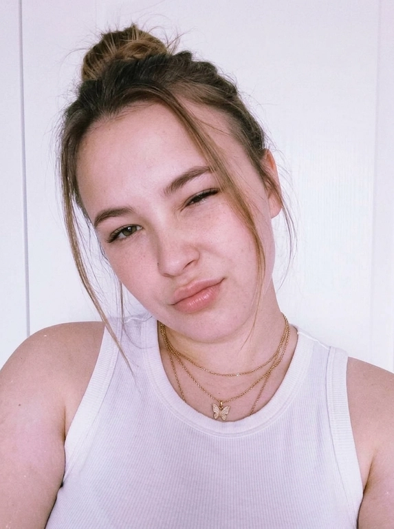 Vertical close-up portrait in the style of a natural selfie: a woman with her head tilted and a soft playful expression, one eye squinted or closed, gaze directed at the camera.
Expressive full lips, neat eyebrows, and soft makeup with an emphasis on the lips and eyes are noticeable on the face. There are thin chains around her neck, one with a small butterfly pendant.
The clothes are a white ribbed tank top with thin straps, revealing the shoulders and upper chest. The background is almost completely white, empty and minimalistic, like a light wall, without unnecessary objects.
The light is very soft, diffused, slightly overexposed, with a cool pink and purple hue, creating a gentle, slightly dreamy atmosphere.
The composition is close and intimate, the frame is cropped around the shoulders and chest, with emphasis on the face. The camera is at face level, creating the feeling of taking a picture from a phone. Light graininess, a gentle loss of detail in the highlights, natural, fashionable, slightly vintage aesthetic.



Причёска: Hair gathered into a relaxed, messy bun with soft volume at the roots and a few loose strands framing the face without covering the eyes.