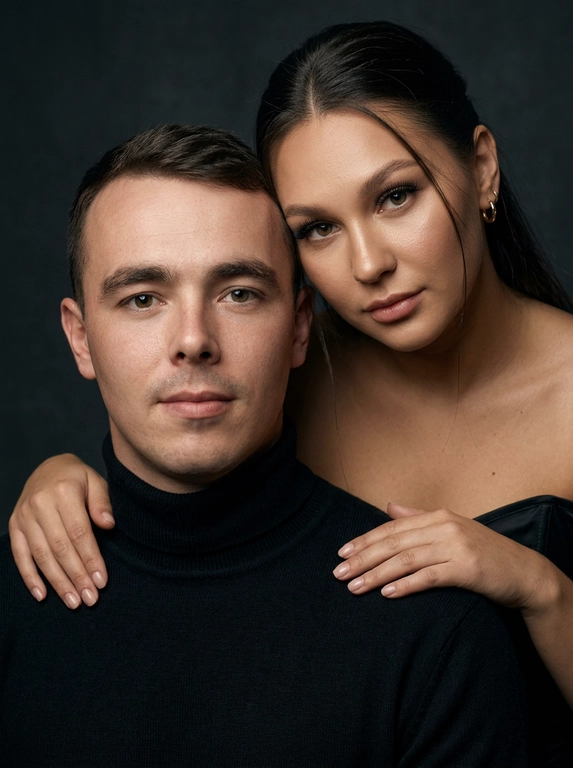 Гиперреалистичный студийный fashion-портрет молодой пары по загруженному референсу, 100% сходство лиц. Черты лица не изменять. Крупный план, кадр по плечи. Мужчина, спокойный уверенный взгляд прямо в камеру, естественное выражение лица, одет в черный трикотажный свитер с высоким воротом. Женщина стоит позади мужчины и нежно обнимает его, ее руки лежат на его плечах. Ее лицо расположено рядом с его лицом, она повернута слегка боком, голова немного наклонена вперед, взгляд направлен прямо в камеру. У женщины выразительные глаза с длинными густыми черными ресницами, естественный clean-glam макияж, натуральные брови, теплый оливковый оттенок кожи, небольшие серьги-кольца. Композиция плотная и интимная, их лица расположены очень близко друг к другу, современный модный портрет пары. Женщина одета в черный корсет. Кожа максимально натуральная: живая текстура кожи, видимые микропоры, subtle skin glow, естественный мягкий блеск кожи, natural skin microtexture, skin translucency, micro skin details, легкие естественные несовершенства кожи, без ретуши и без эффекта пластика. Освещение: мягкий студийный beauty lighting, рассеянный ключевой свет спереди и немного сбоку, мягкие глубокие тени для объема лица, деликатные световые блики на коже. Фон: глубокий темный студийный фон, темно-графитовый или почти черный, создающий контраст с кожей и лицами. Стиль: luxury fashion editorial photography, high fashion portrait, ultra photorealistic, 85mm portrait lens, shallow depth of field, creamy bokeh, cinematic soft lighting, RAW photo, extremely detailed, fine hair strands, natural skin pores, professional photography, photorealism, 8k, high dynamic range.