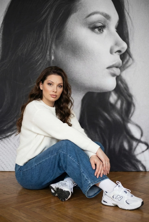   Максимально сохранить внешность, причёску и длину, как на фото.
Женщина должна сидеть на полу в изящной позе, в современном белом джемпере , в широких джинсах и белых носках и белых  кроссовках New Balance.

Её волосы как на фото
Выражение лица, как на фото, она смотрит на камеру .

Фон — тот же её профиль, в мягком крупном чёрно-белом художественном стиле, крупный план сбоку.

Композиция должна быть художественной, мягкой, реалистичной.
Создай изображение, которое выглядит как настоящая фотография.
